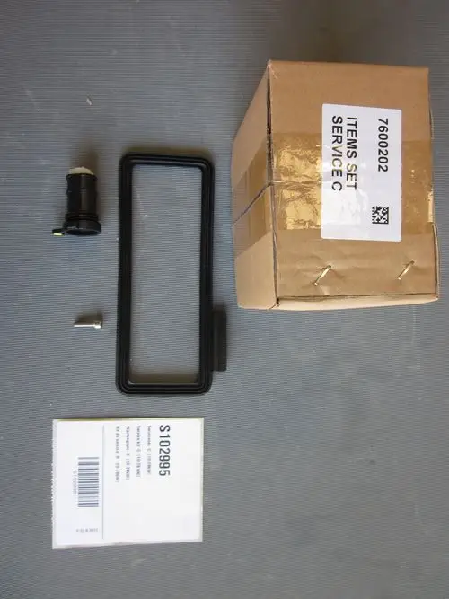 Remeha-Serviceartikel auf einem Untergrund: ein schwarzer Schlauchanschluss, ein rechteckiger Rahmen, eine Schraube und eine beschriftete Verpackung.